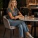 Blonde woman in a gray T-shirt and jeans sitting alone at a modern bar table