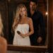 Blonde woman wrapped in a white towel brushing wet hair at a bathroom mirror with a dark-haired man behind her