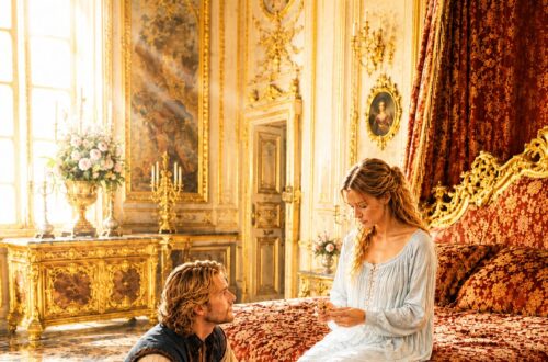 King Mark kneeling beside Lili as she sits on the bed in a royal bedroom