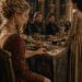 King Mark leaning toward seated Lili as he tries to see her face