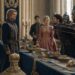 Lili standing between two men beside a banquet table in a crowded royal hall