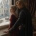 Blond man embracing a woman in a burgundy gown beside a palace window