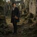 Aiden standing among the overgrown stone ruins of an old castle with a book in his hand