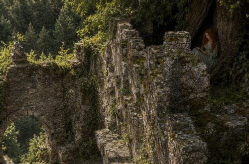 Aiden looking up at Tess as she sits reading beneath a tree above the castle ruins