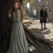 Nervous Tess standing against an old tree while Aiden watches her among the castle ruins
