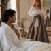 Tess struggling to fasten a long skirt while Aiden watches from their bed