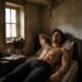 Shirtless Aiden lying on a simple bed in a rustic room beside a candle and glass of wine