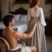 Shirtless Aiden fastening the back of Tess's long skirt as she stands beside their bed