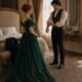 Tess in an emerald green gown facing Aiden as he rolls up his sleeves in their bedroom