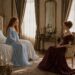 Tess in a pale blue dress speaking privately with an elegant red-haired woman in burgundy