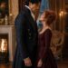 Aiden standing face to face with a red-haired woman in burgundy beside a fireplace