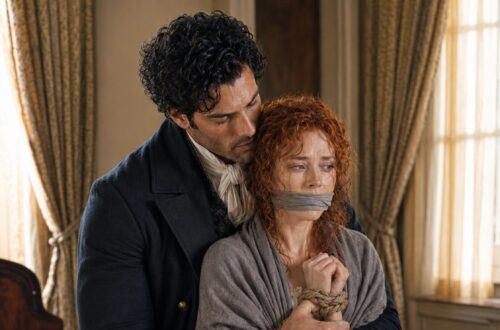 Aiden holding bound and gagged Tess on a bed as she sits wrapped in a gray cloak