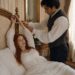 Aiden tying Tess's bound wrists to the carved headboard while she lies on the bed