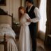 Aiden supporting a weak Tess by the arm as she stands beside their bed in a white nightgown