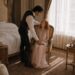 Aiden leaning over seated Tess and placing his hand over hers and the book in her lap
