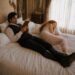 Aiden reading on the bed while Tess sleeps peacefully beside him in a soft pink nightgown